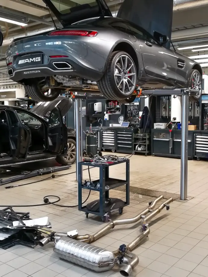 Kuvassa AMG mersu nosturilla ylös nostettuna ja konepelti auki. Maassa kiiltävä tupla-pakoputkisto, ja auton alla työkalukärry täynnä osia ja työkaluja.