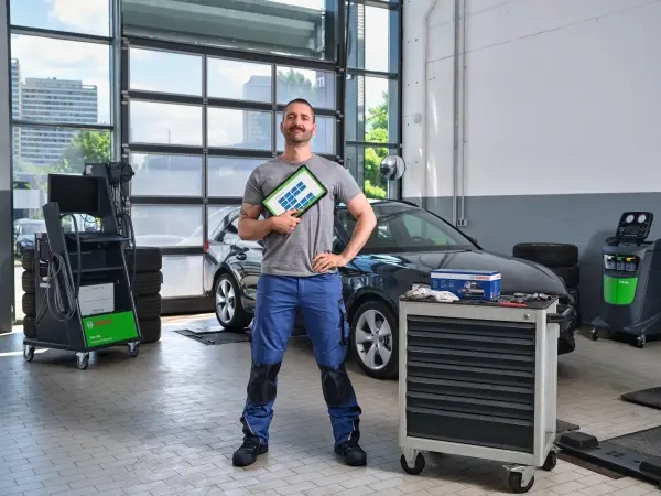 Mies seisoo korjaamohallissa kädessään tablettitietokone, jossa ESI Evolution ohjemisto. Taustalla henkilöauto, ja muita Bosch laitteita kuten FSA-testeri ja ilmastointihuoltolaite.